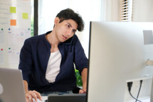 Young Man with dark hair talking on phone in front of silver computer 